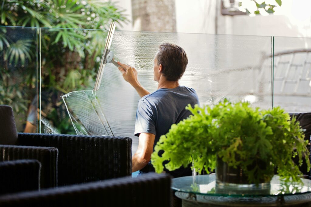 Man in a gray shirt cleaning a clear glass wall using a squeegee.