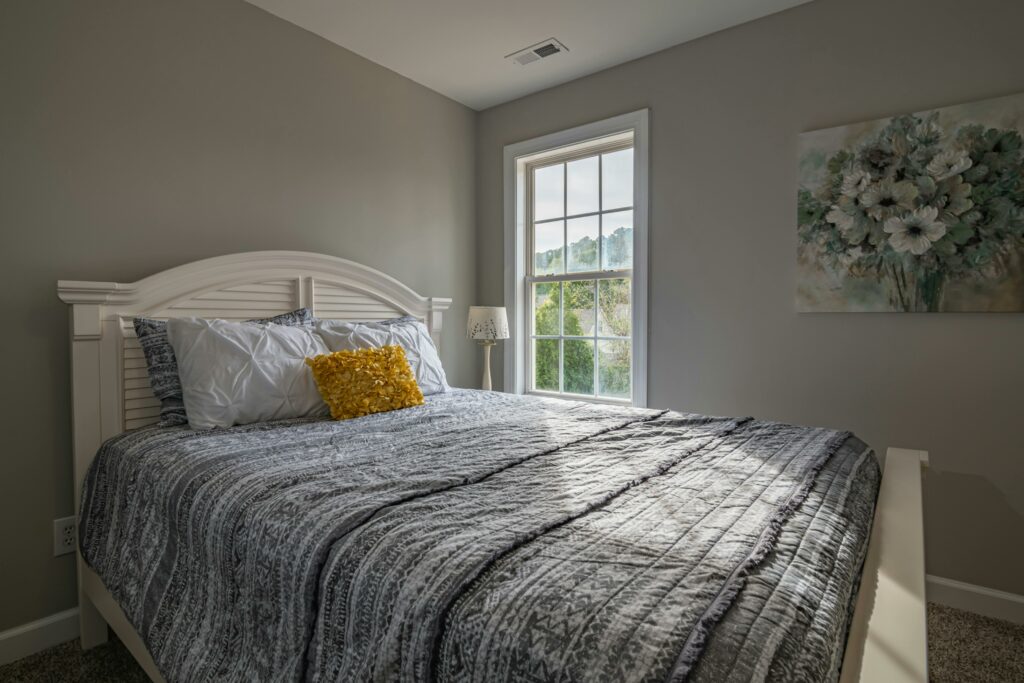 Bright, neatly made bed with patterned bedding and a yellow accent pillow in a cozy, sunlit bedroom with neutral walls and a large window.