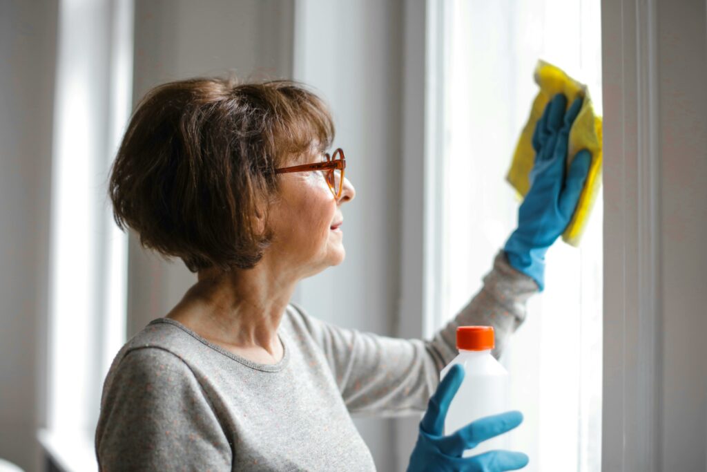 Woman wearing gloves and glasses cleaning a window with a yellow cloth and cleaning solution, focusing on maintaining a clean and bright home environment.