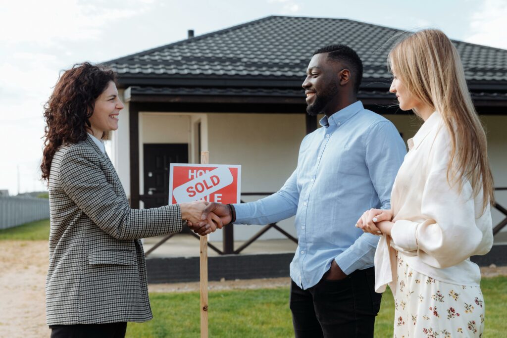 Real estate agent shaking hands with a smiling couple in front of a house with a 'Sold' sign, symbolizing a successful home sale.