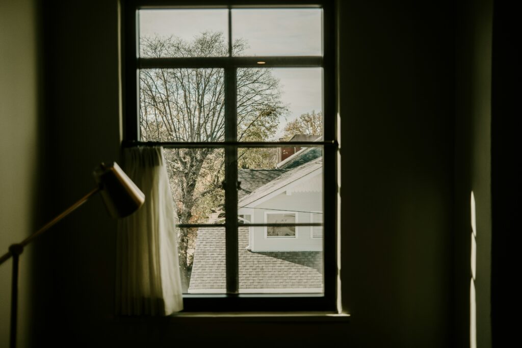 View through a window from a dimly lit room, showing a house roof and leafless trees outside on a clear day, with a floor lamp and curtain in the foreground.