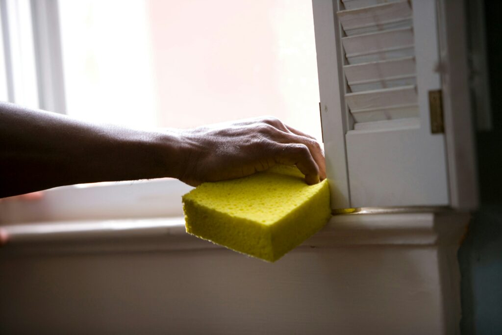 Close-up of a person cleaning a window sill with a yellow sponge near white shutters in natural daylight.