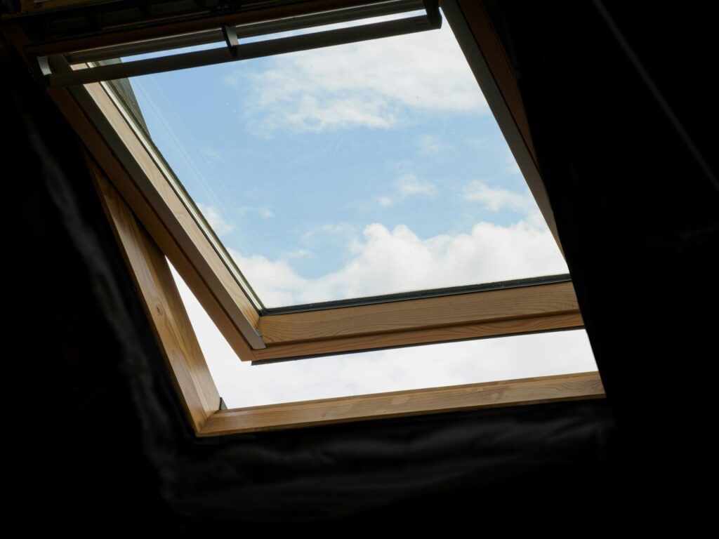 View of an open skylight window framed in wood, looking up at a blue sky with scattered white clouds.