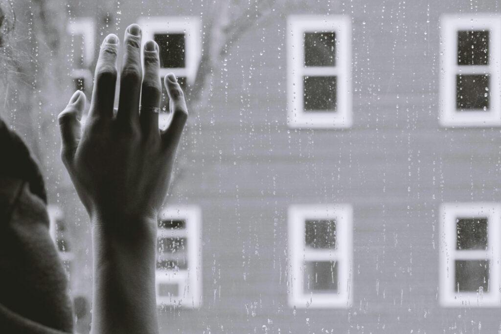 Hand touching a rain-covered window with blurred view of a building in the background on a gloomy day