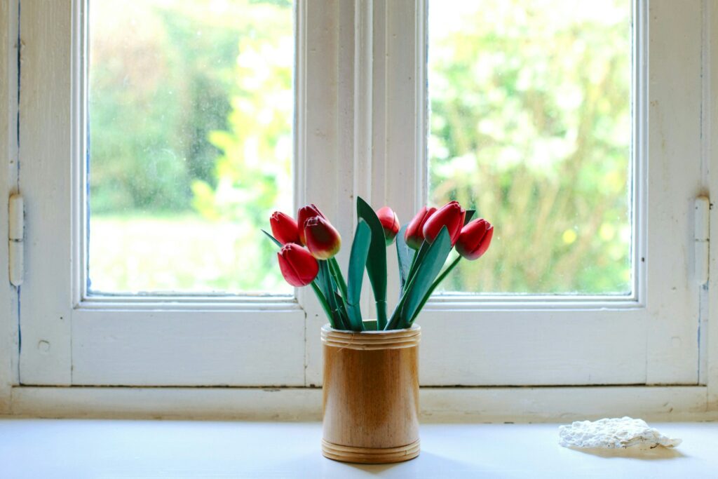 Bright window with slightly hazy glass, a wooden vase holding red tulips on the windowsill, and soft greenery visible outside.