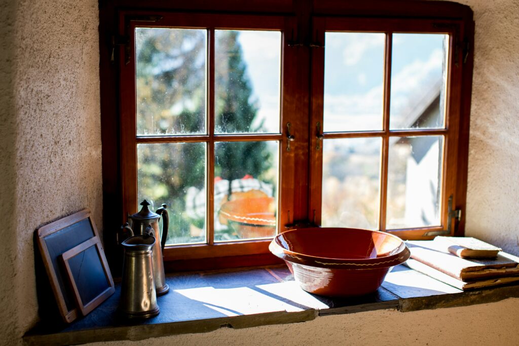 Rustic kitchen window with wooden frames and slightly dirty glass, showing sunlight streaming in over vintage pitchers, a red ceramic bowl, and stacked books on the windowsill.
