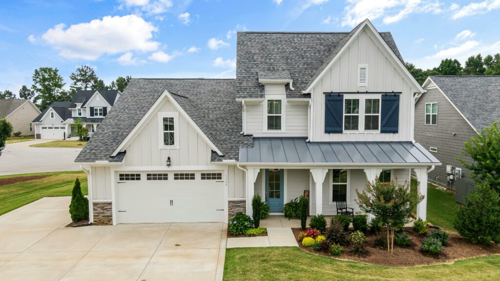 Modern farmhouse-style home with white vertical siding, dark blue shutters, a gray shingle roof, and a covered front porch, surrounded by neatly landscaped greenery and a spacious concrete driveway.