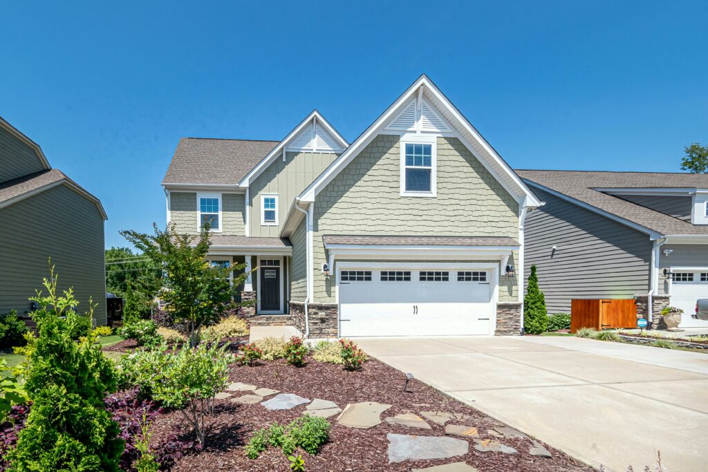 A modern two-story suburban house with green siding, white trim, and a two-car garage. The home features stone accents on the lower facade and a gabled roof. A landscaped front yard with mulch, decorative plants, shrubs, and a stone walkway enhances curb appeal. The driveway is wide and clean, and the sky is clear with bright sunlight, indicating a pleasant day.