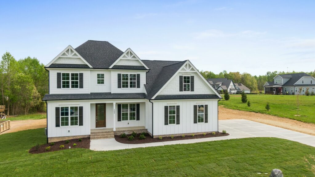 A newly constructed two-story white house with black shutters and a dark gray roof, featuring a covered front porch, fresh landscaping, and a spacious concrete driveway set in a suburban neighborhood with open green space and trees in the background.