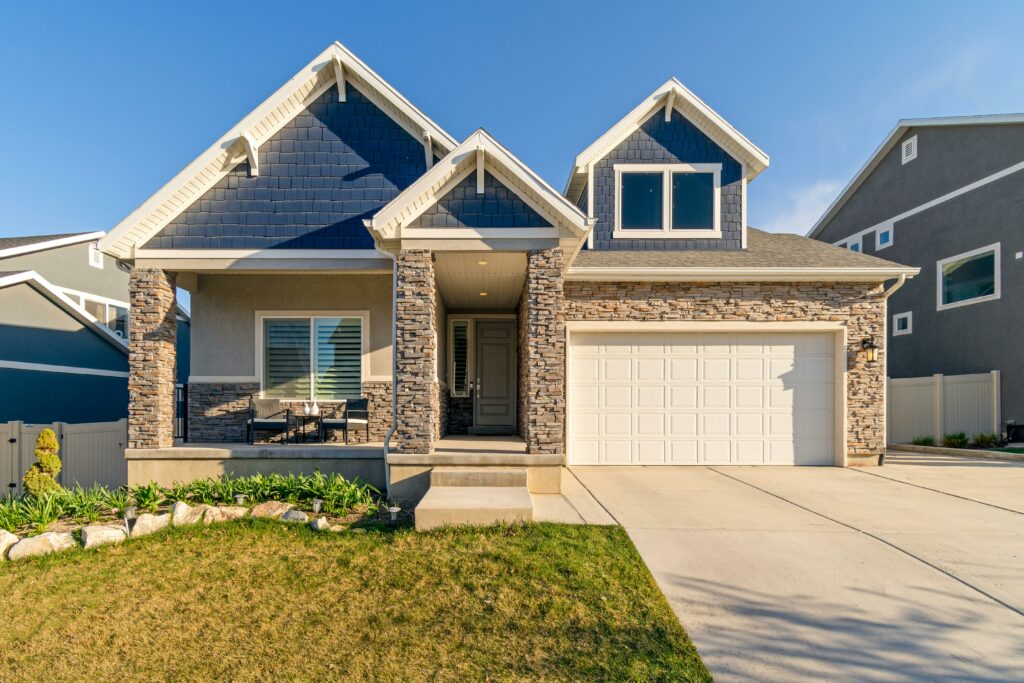 A modern suburban house with a stone and siding exterior, featuring a two-car garage, gabled roof, and a small covered front porch with outdoor seating. The home has well-maintained landscaping with green plants along the front and a wide concrete driveway. The sky is clear and blue, suggesting a bright, sunny day.