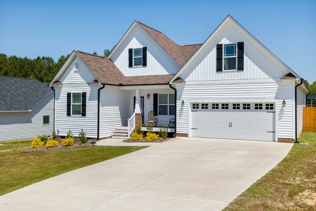 A single-family white house with black shutters and a brown roof, featuring a two-car garage, a small covered front porch with outdoor seating, and a wide concrete driveway leading to a neatly landscaped front yard.