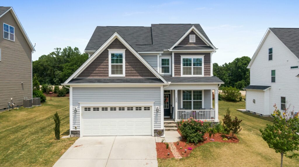 Two-story suburban house with a gray and white exterior, dark shingle roof, attached two-car garage, and a neatly landscaped front yard with flowers and shrubs.