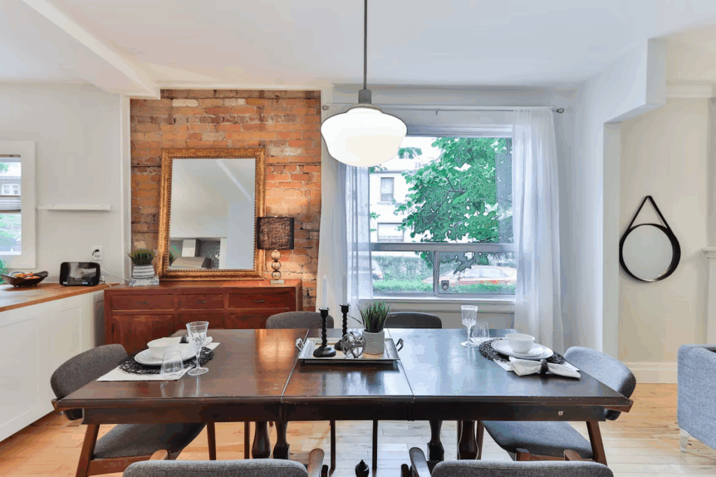 A stylish dining room with a dark wooden table set for six, featuring modern gray chairs and simple tableware. The room has a mix of rustic and contemporary design elements, including an exposed brick accent wall with a large framed mirror, a white pendant light overhead, and sheer curtains framing a large window. Natural light fills the space, highlighting the cozy, inviting atmosphere.
