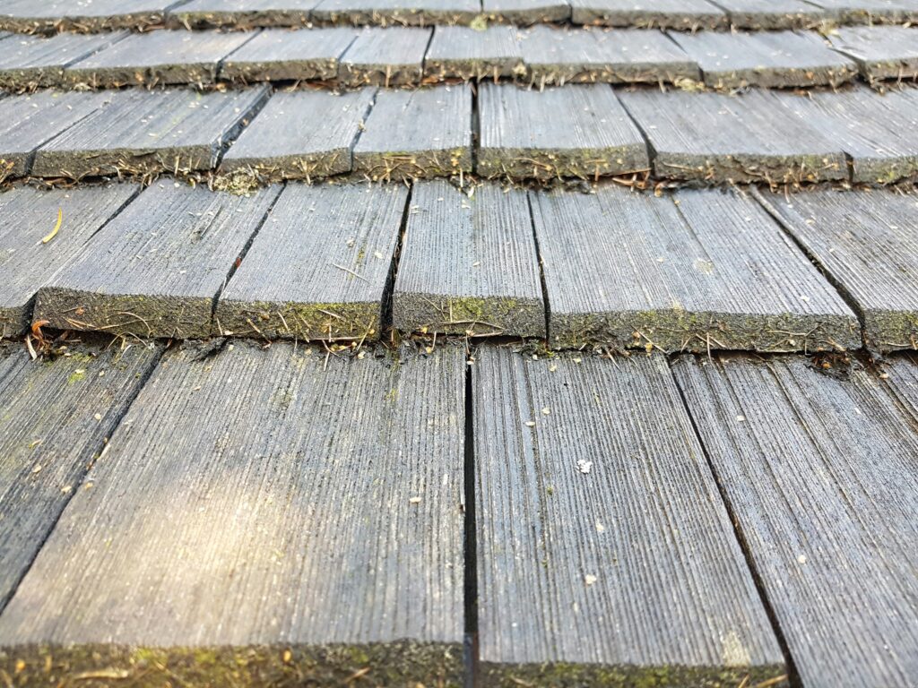 Close-up of weathered wooden roof shingles with visible gaps, dirt, and moss growth along the edges. 