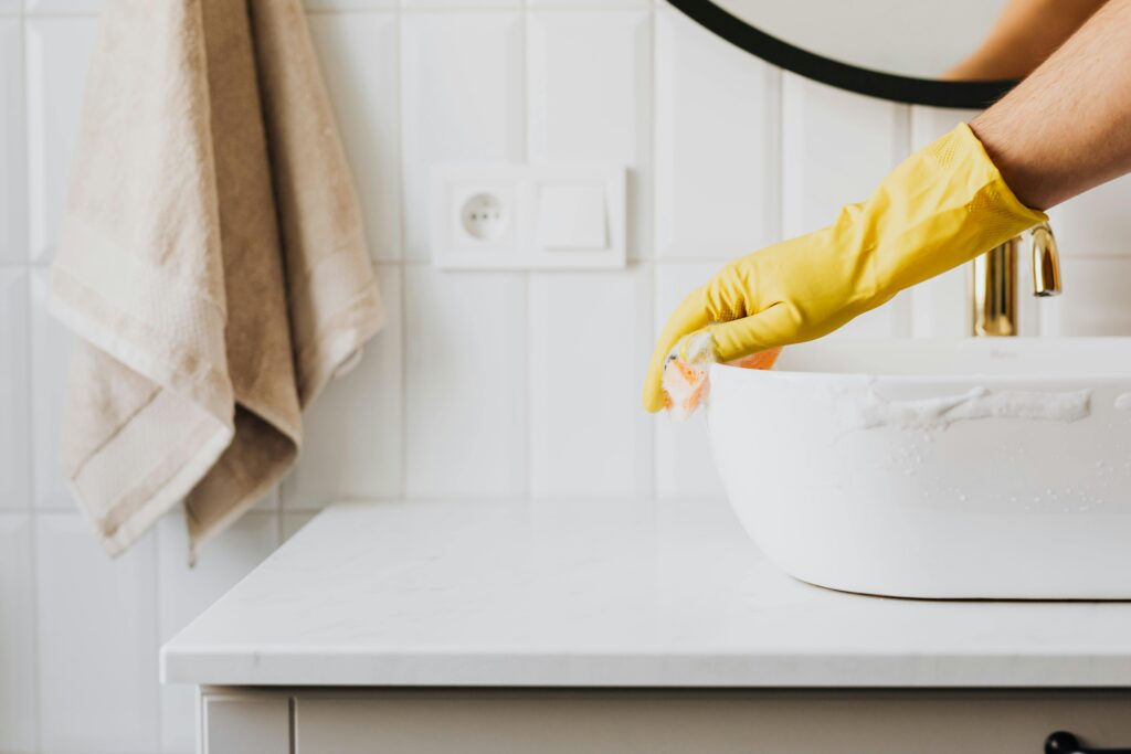Person wearing yellow rubber gloves cleaning a white bathroom sink with a sponge, next to a hanging beige towel.