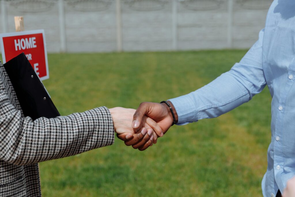 Close-up of two people shaking hands outdoors in front of a home for sale sign, symbolizing a real estate deal or agreement.