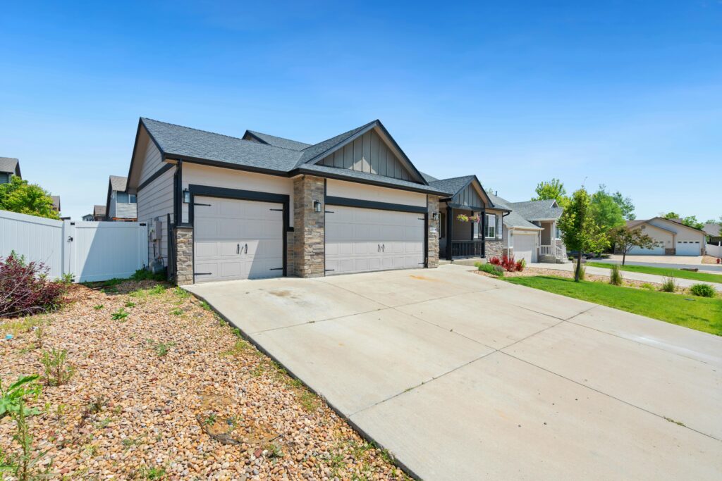 Modern single-story house with a large concrete driveway, stone and siding exterior, double garage doors, and neatly landscaped front yard under a bright blue sky.