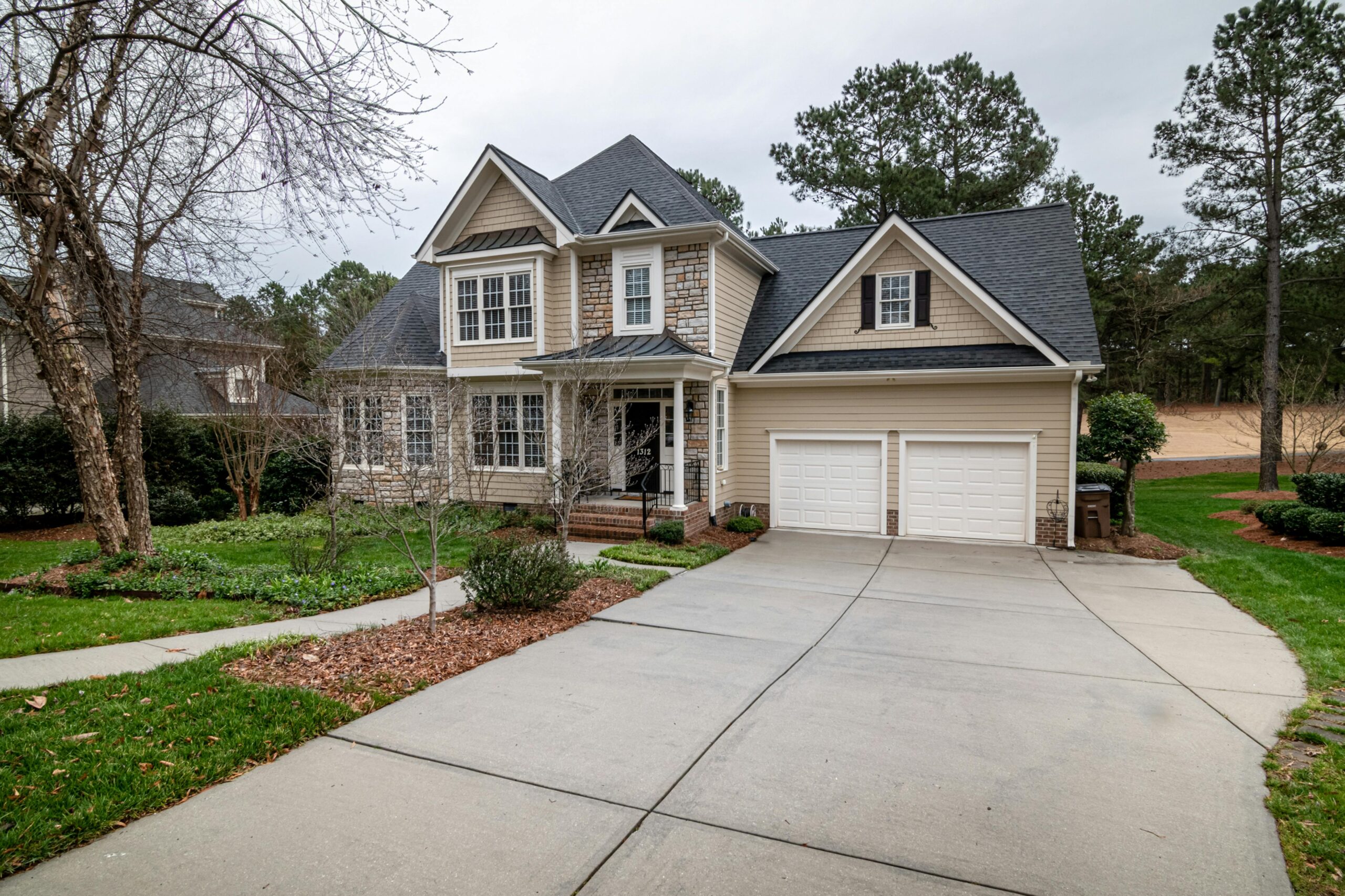Front view of a modern two-story suburban house with beige siding, stone accents, and a double garage, featuring a clean concrete driveway and neatly landscaped yard with trees and shrubs.