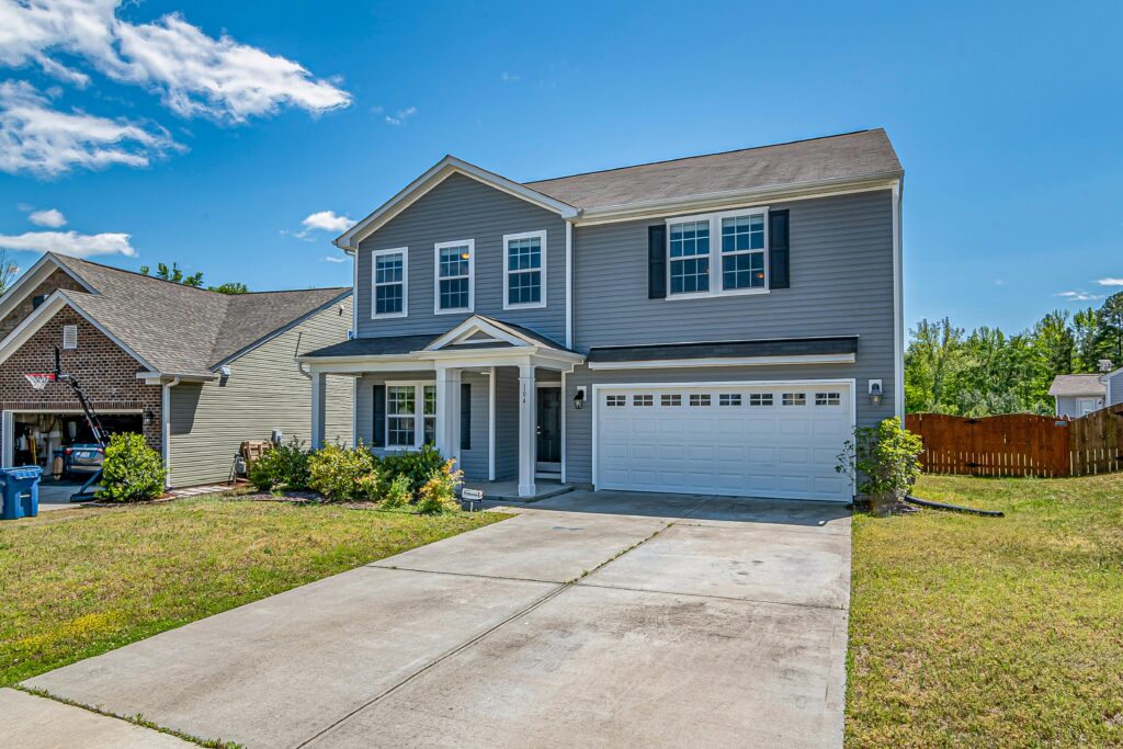 Two-story gray house with white trim and a double garage, featuring a clean concrete driveway, small front porch, and neatly maintained yard under a bright blue sky.
