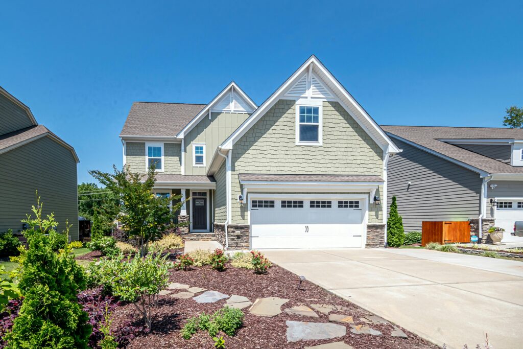 Two-story light green house with white trim and stone accents, featuring a double garage, clean concrete driveway, and landscaped front yard with shrubs and flowers under a clear blue sky.
