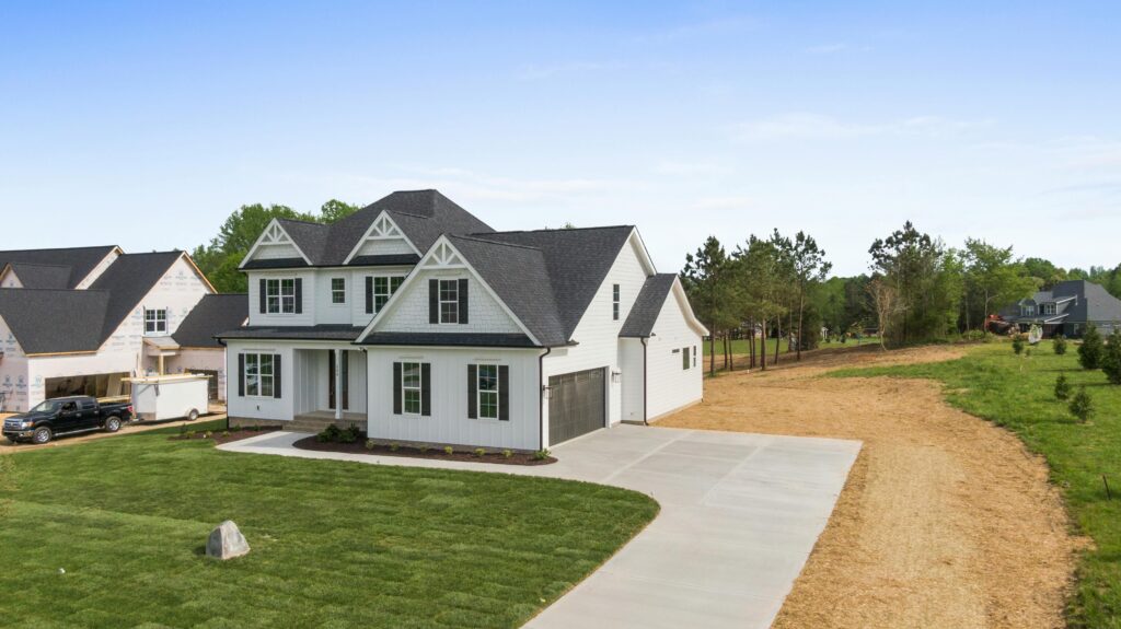 Modern two-story white farmhouse-style home with dark roof and black garage door, featuring a wide concrete driveway, freshly landscaped lawn, and neighboring new construction homes in a semi-rural setting.
