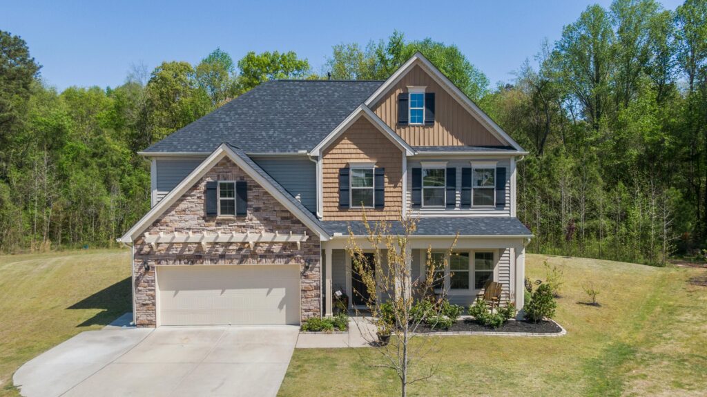 A two-story suburban house with a combination of stone, siding, and shingle exterior, featuring a two-car garage, black shutters, and a well-kept front yard with young trees and landscaped garden beds, set against a backdrop of tall green trees.