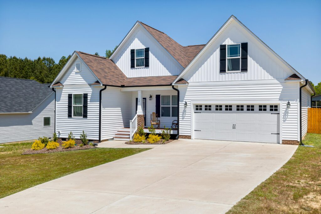 A modern suburban white house with dark shutters, a brown shingle roof, and a clean driveway under a clear blue sky, featuring well-maintained landscaping and a covered front porch with outdoor seating.