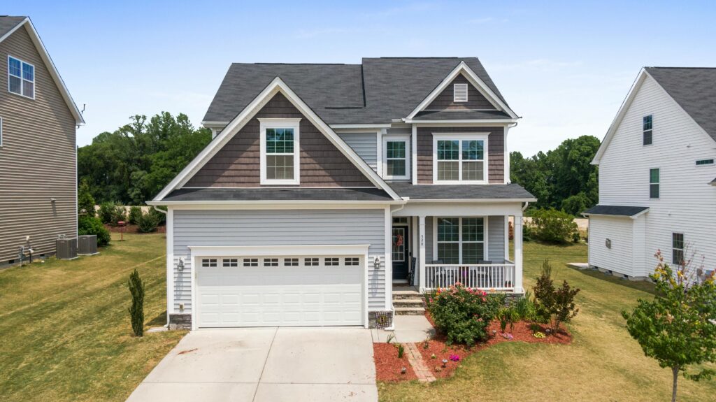 Two-story suburban house with gray siding, white trim, and dark shingle accents, featuring a white double garage, front porch, and neatly landscaped yard on a clear day.