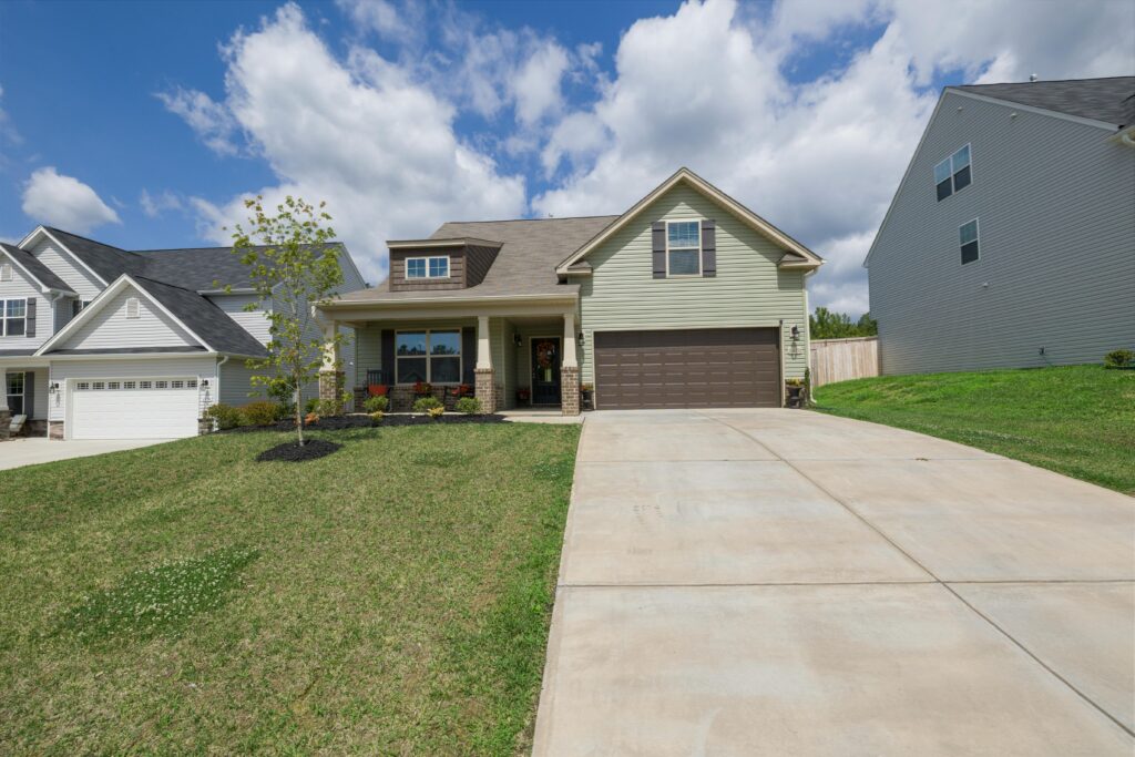 Front view of a single-story suburban home with green siding and a dark brown garage door, featuring a wide concrete driveway, neatly trimmed lawn, and a small landscaped area with a young tree and shrubs.
