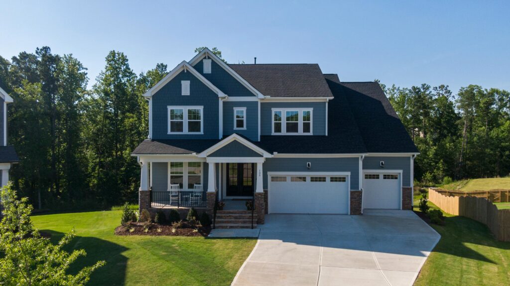 A two-story blue suburban house with white trim, black roof shingles, and a three-car garage, featuring a covered front porch with rocking chairs and surrounded by a neatly landscaped lawn and wooded backdrop.